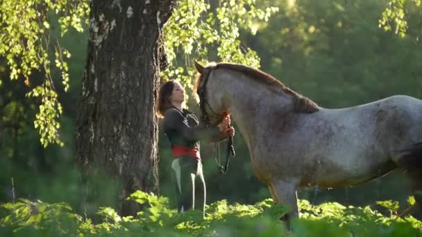 Flicka rider står med en häst nära ett träd mot en grön skog och strålar morgonsolen — Stockvideo