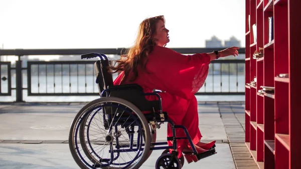 Schöne rothaarige Mädchen im Rollstuhl nimmt aus dem Regal Buch — Stockfoto