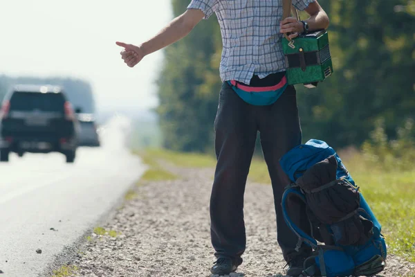 Чоловік у сорочці, темні штани, з акордеоном на плечах, що стоїть біля дороги і ходіння, рюкзак лежить біля ніг — стокове фото