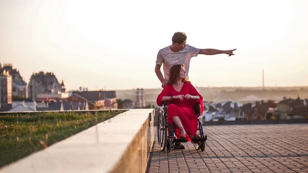 Guy rolls his girlfriend in a wheelchair and shows her something with his hand in the distance, close up