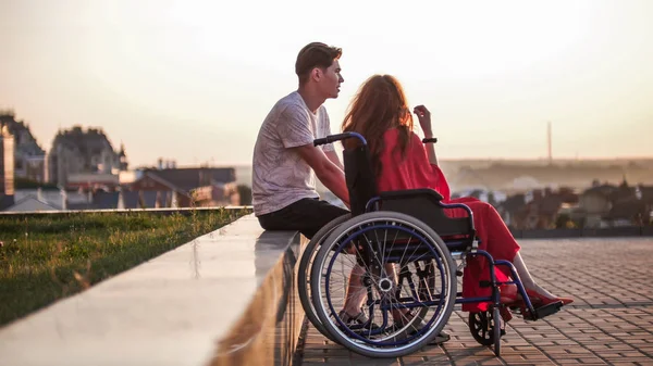 Молода пара захоплюється заходом сонця, дівчина в інвалідному візку, хлопець сидить поруч — стокове фото