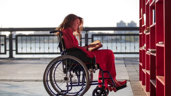 Ein junges schönes Mädchen mit roten Haaren in einem roten Kleid sitzt im Rollstuhl und liest ein Buch — Stockfoto