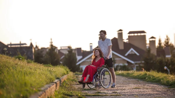 Una bella ragazza su una sedia a rotelle e il suo giovane uomo sono a piedi nel parco, accanto a belle case — Foto Stock