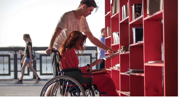 Молодий чоловік показує книжки своїй дівчині з рудим волоссям, яка сидить у інвалідному візку — стокове фото