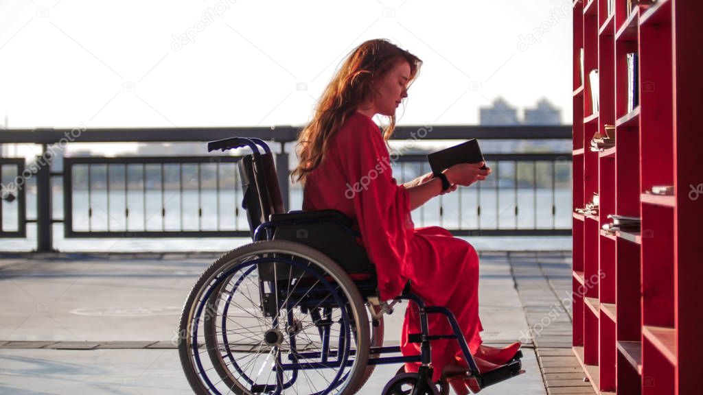 A young girl with red hair in a red dress is sitting in a wheelchair and reading a book