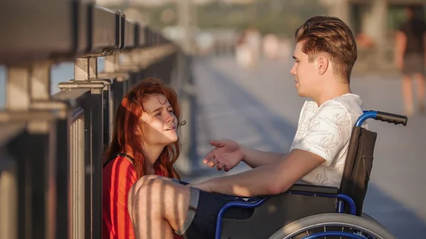 Der Mann im Rollstuhl sagt etwas zu dem rothaarigen Mädchen und sie schaut vorsichtig zu, an einem Sommerabend auf der Promenade. — Stockfoto