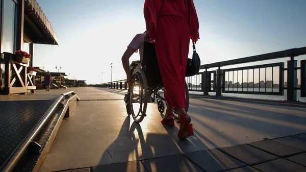 Ein Mädchen mit roten Haaren in orangefarbenem Kleid wälzt einen behinderten Mann im Rollstuhl auf der Uferpromenade im Sonnenuntergang — Stockfoto