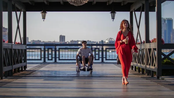Красива дівчина з рудим волоссям у червоній сукні йде вздовж набережної і за нею молодий хлопець їде в інвалідному візку — стокове фото