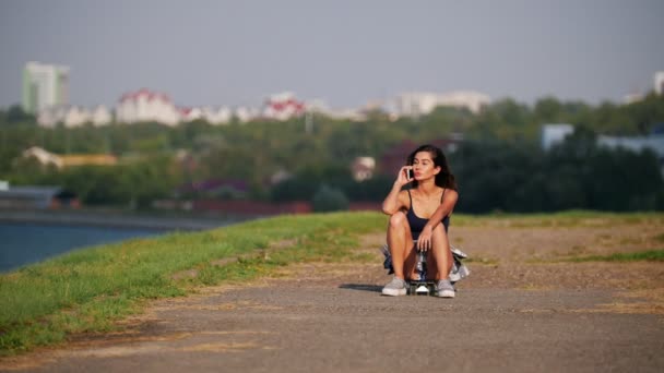 Krásná mladá dívka sedí na skateboardu a mluví po telefonu na pobřeží — Stock video