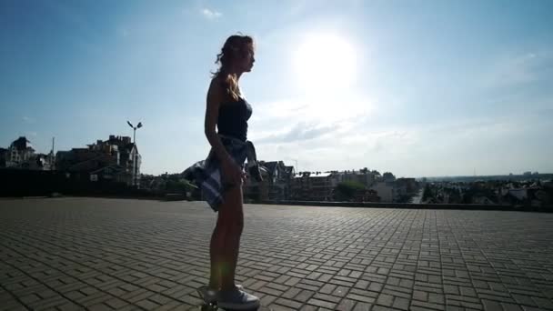Девушка катается на скейтборде на пустой площади на фоне прекрасного вида на город — стоковое видео