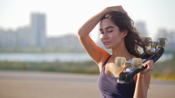 Una hermosa chica con una mano sosteniendo un patín, la otra tira de su pelo, mira a la cámara — Vídeo de stock