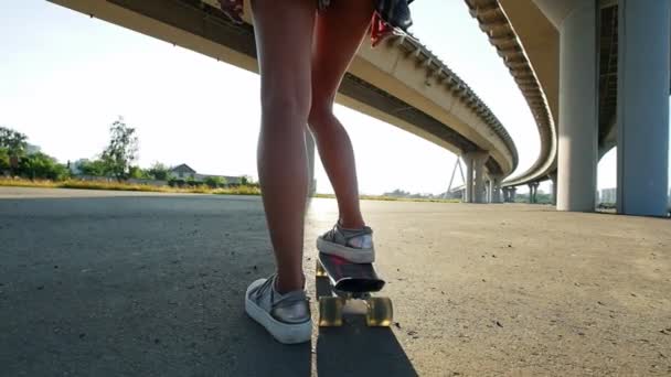 Chica joven con piernas delgadas montando un monopatín debajo del puente — Vídeo de stock