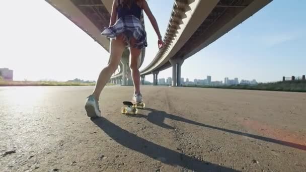 Menina esbelta em shorts curtos e uma camisa xadrez em seu cinto monta um skate sob a ponte — Vídeo de Stock