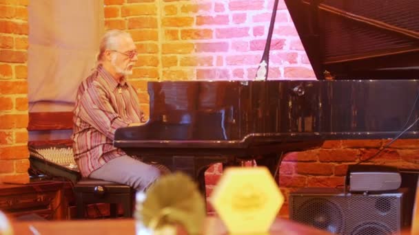 Harmaahiuksinen mies, jolla on häntä päässä ja lasit ja joka soittaa pianoa jazz-baarissa. — kuvapankkivideo