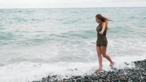 Jong meisje wandelen langs de rotsachtige kust van de zee. Er zijn golven. De lange haren van meisjes is opgeblazen door de wind. — Stockvideo