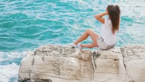 Szép, fiatal lány ül a sziklák, kiegyenesedik a haját, és élvezi egy frissítő fuvallat és egy gyönyörű kilátással a tengerre — Stock videók