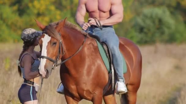 Un joven con un físico fuerte sentado en un caballo a caballo, una joven instructora cuenta y muestra cómo mover a un hombre a caballo — Vídeos de Stock