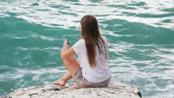 Hermosa joven se sienta en las rocas, endereza su cabello y disfruta de una brisa refrescante y una hermosa vista del mar — Vídeos de Stock