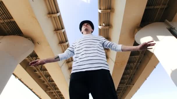 Ein junger Mann mit Hut und gestreiftem T-Shirt, der an einem heißen Sommertag unter der Brücke tanzt — Stockvideo