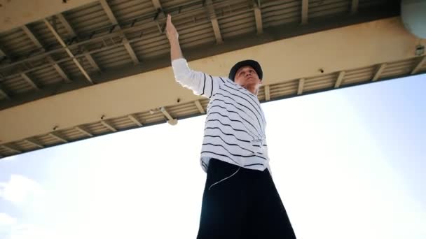 Un jeune danseur de rue dans un chapeau se déplace vers la musique avec des écouteurs sous le pont un jour d'été — Video