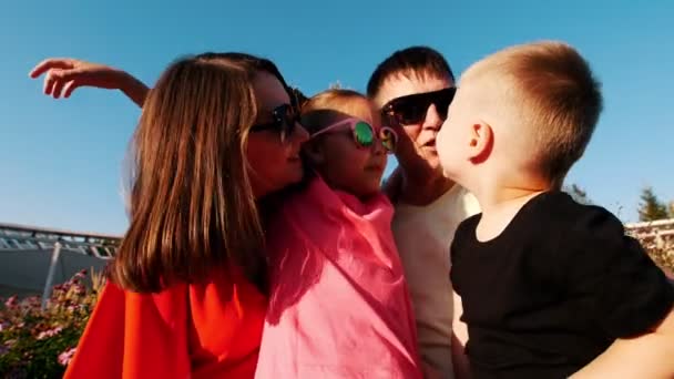 Glückliche Familie im Park auf einer Bank sitzend. kleiner sohn küssen papa, mama und ältere schwester. — Stockvideo