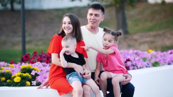 Una familia joven, que consta de 4 personas, se sienta en el parque junto a las hermosas flores . — Vídeo de stock