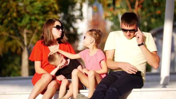 Una joven familia sentada en el parque bajo el sol. Papás al teléfono. . — Vídeos de Stock