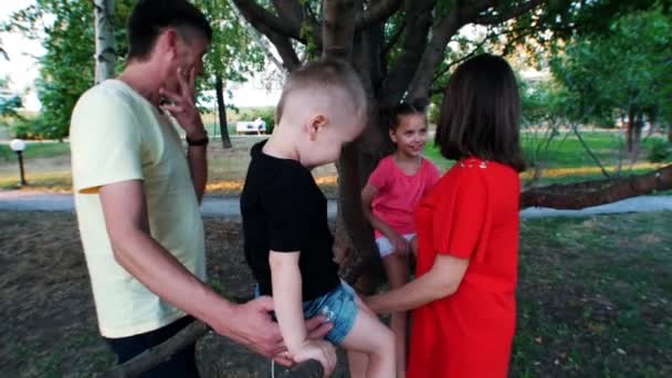 Vierköpfige Familie steht neben einem Baum. Kinder sitzen auf großen Ästen, Eltern halten Kinder. — Stockvideo