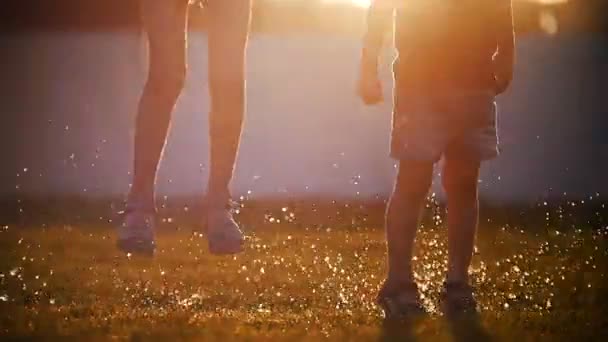 Chica y niño saltando sobre charcos en la hierba. Aerosol volador que brilla al sol . — Vídeos de Stock