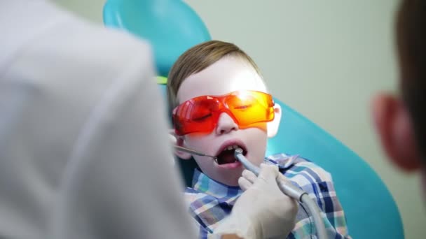 Детский дантист лечит мальчика, сидящего в кресле в защитных очках оранжевого цвета — стоковое видео