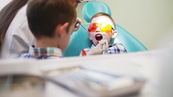 La mujer dentista conduce una herramienta en la boca de un niño paciente. El hermano de los chicos está cerca. . — Vídeos de Stock