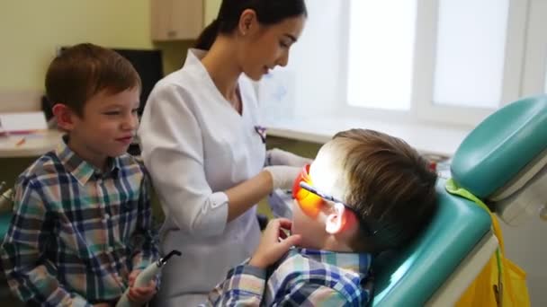 O dentista prepara as ferramentas para o tratamento. O rapaz paciente está à espera na cadeira. O outro rapaz, o irmão gémeo, aplaude o irmão. . — Vídeo de Stock