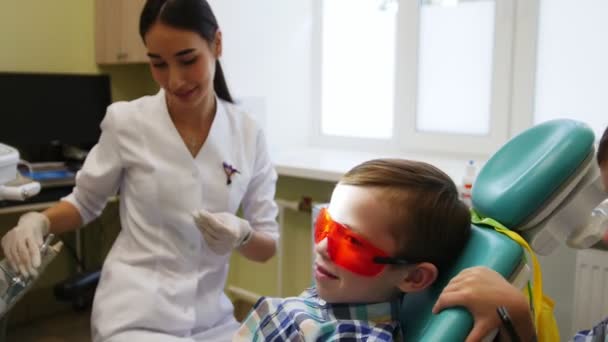 El joven dentista se prepara para una sesión de tratamiento. Un pequeño paciente se sienta en una silla y espera. Su hermano gemelo lo divierte. . — Vídeos de Stock