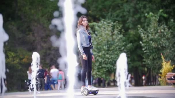 Bella ragazza snella con i capelli lunghi cavalca un giroscopio nel parco passato fontane e persone, lancia uno sguardo alla fotocamera — Video Stock