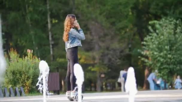 Молодая девушка ездит на гироскопе и разговаривает по телефону в парке в летний день, в фонтанах на переднем плане — стоковое видео
