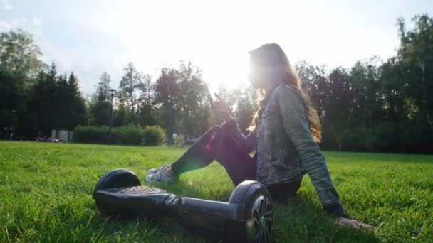 Linda joven sentada en la hierba en el parque, sosteniendo el teléfono en sus manos, correspondencia. El giroscopio está junto a sus pies. . — Vídeos de Stock