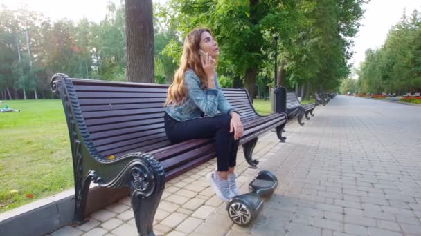 Красивая девушка сидит на скамейке и разговаривает по телефону. Рядом с ее ногами гироскоп. . — стоковое видео