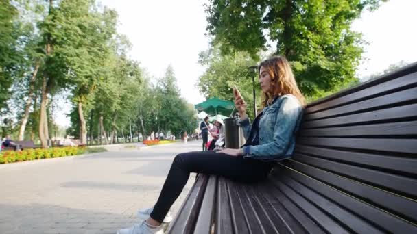 Ein junges Mädchen sitzt mit einem Handy auf einer Bank im Park, in der Nähe der Füße steht ein Kreisel. — Stockvideo