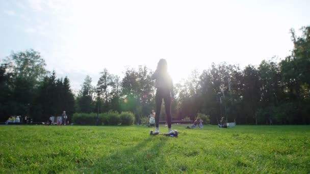 Ein junges Mädchen mit einer schönen Figur reitet auf einem Kreisel auf dem Gras im Park. die Sonne scheint. — Stockvideo