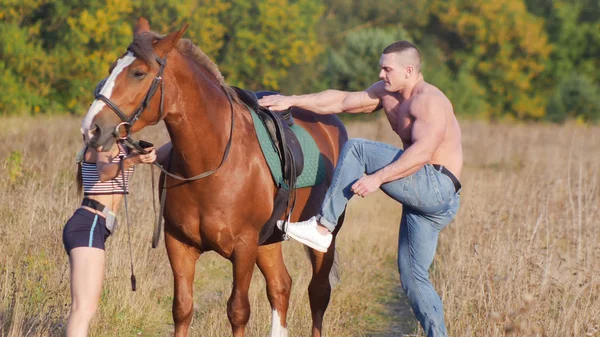 Jovem com um físico forte sobe em um cavalo, uma jovem mulher esbelta segurando um cavalo — Fotografia de Stock