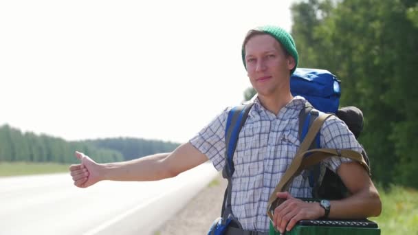 Un hombre joven con una camisa a cuadros, un sombrero verde y una mochila de pie junto a la carretera y los coches de bocina — Vídeos de Stock