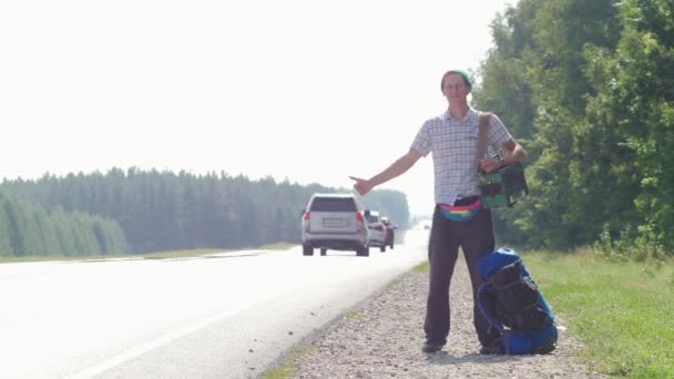 Um homem com uma camisa xadrez, um chapéu azul, com um acordeão no ombro ao pé da estrada e buzinando carros passando, deitado ao lado da mochila . — Vídeo de Stock