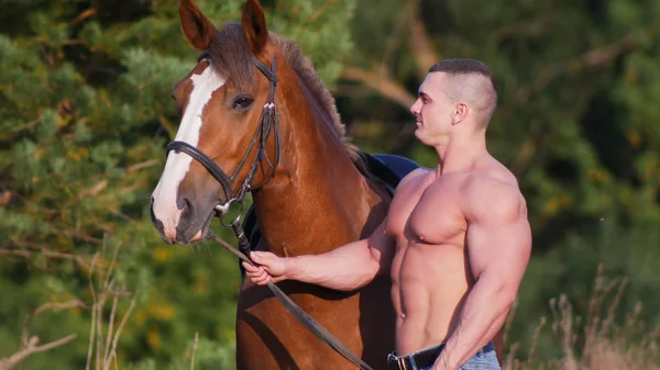 Homem atlético com tronco nu em calças jeans ao lado do cavalo, segurando o cavalo pelo freio — Fotografia de Stock