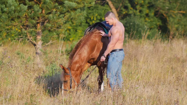 Egy erős férfi meztelen felsőtestét és farmer nadrág áll egy ló, hogy füvet rág — Stock Fotó