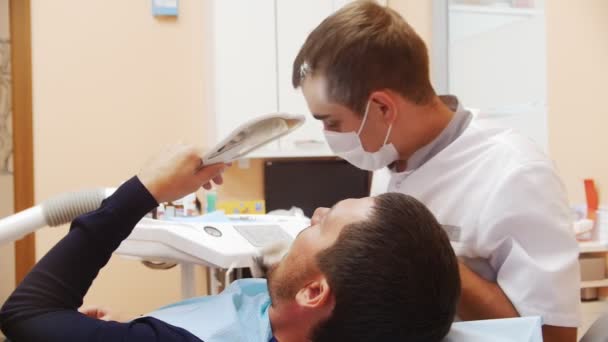 Male dentist in the process of treating a male patients teeth. The patient looks in the mirror. — Stock Video