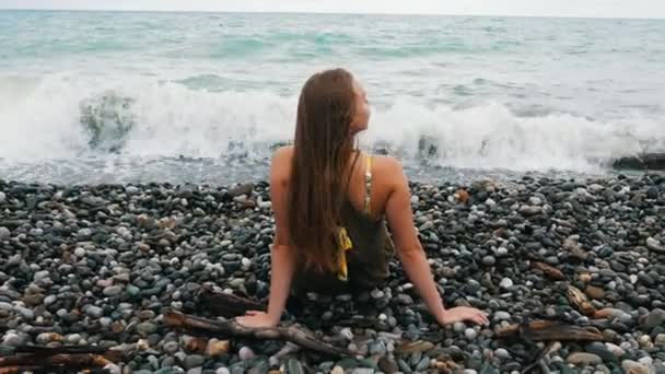 Hermosa joven se sienta en las rocas, endereza su cabello y disfruta de una brisa refrescante y una hermosa vista del mar — Vídeos de Stock