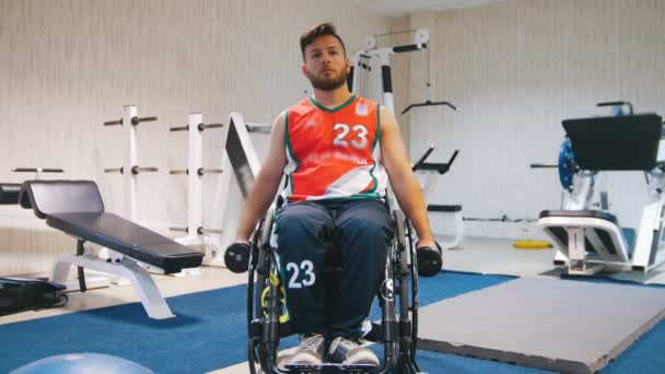 Bonito deficiente em uma cadeira de rodas levanta os braços com halteres no ginásio durante o treinamento . — Vídeo de Stock