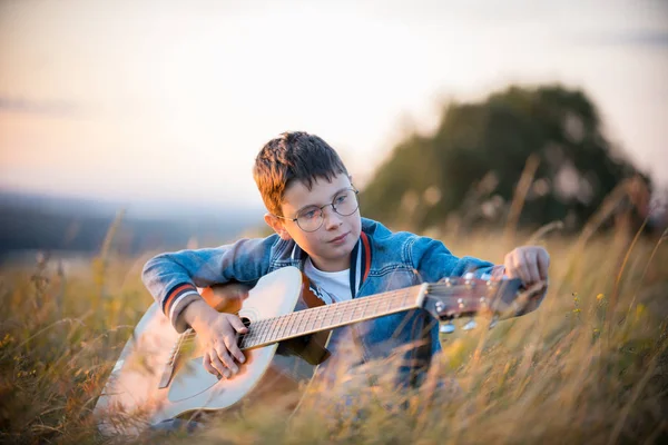 Kleiner Teenie-Junge stimmt Akustikgitarre auf Sommerfeld bei Sonnenuntergang und lächelt — Stockfoto