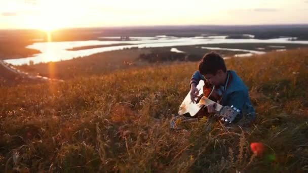 Αγόρι σε στρογγυλά γυαλιά που παίζει κιθάρα στο πεδίο στο ηλιοβασίλεμα — Αρχείο Βίντεο
