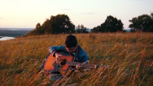 Garçon dans des lunettes rondes jouer de la guitare dans le champ au coucher du soleil — Video